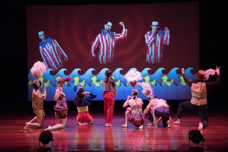 A group of dancers in head-dresses stand on their knee at stare up at a projection that features a man in a Uncle Sam costume.